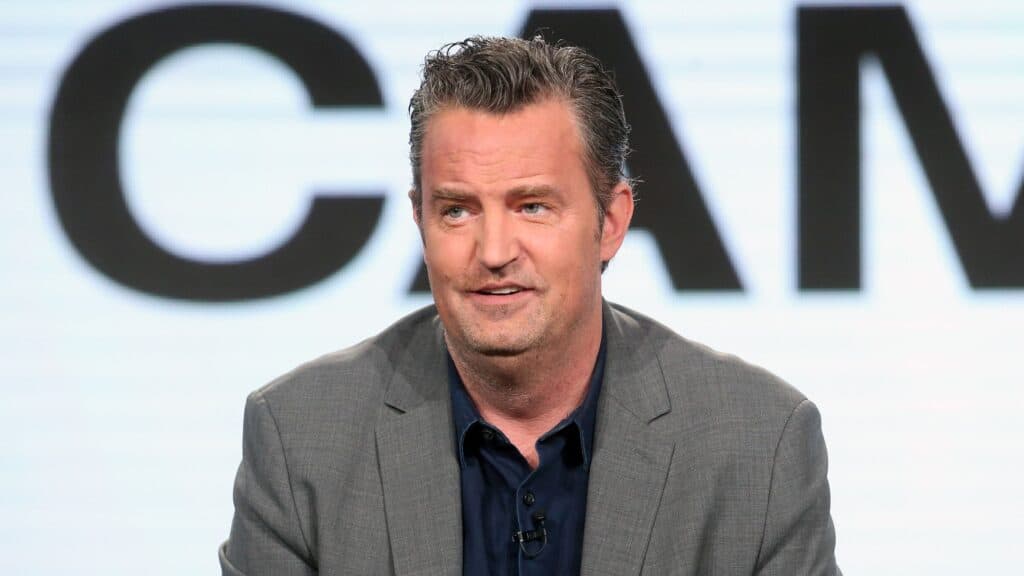 Matthew Perry durante una conferencia, vestido con un traje gris y camisa oscura, mientras habla sobre su carrera en Hollywood.