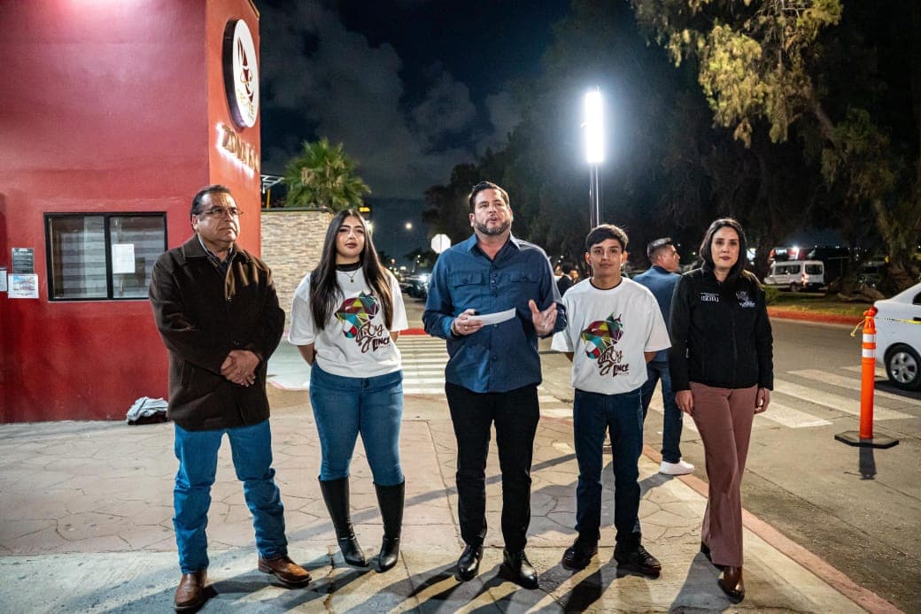 Un grupo de cuatro personas, incluido un hombre que habla, se encuentra frente a un edificio iluminado por la noche, en un