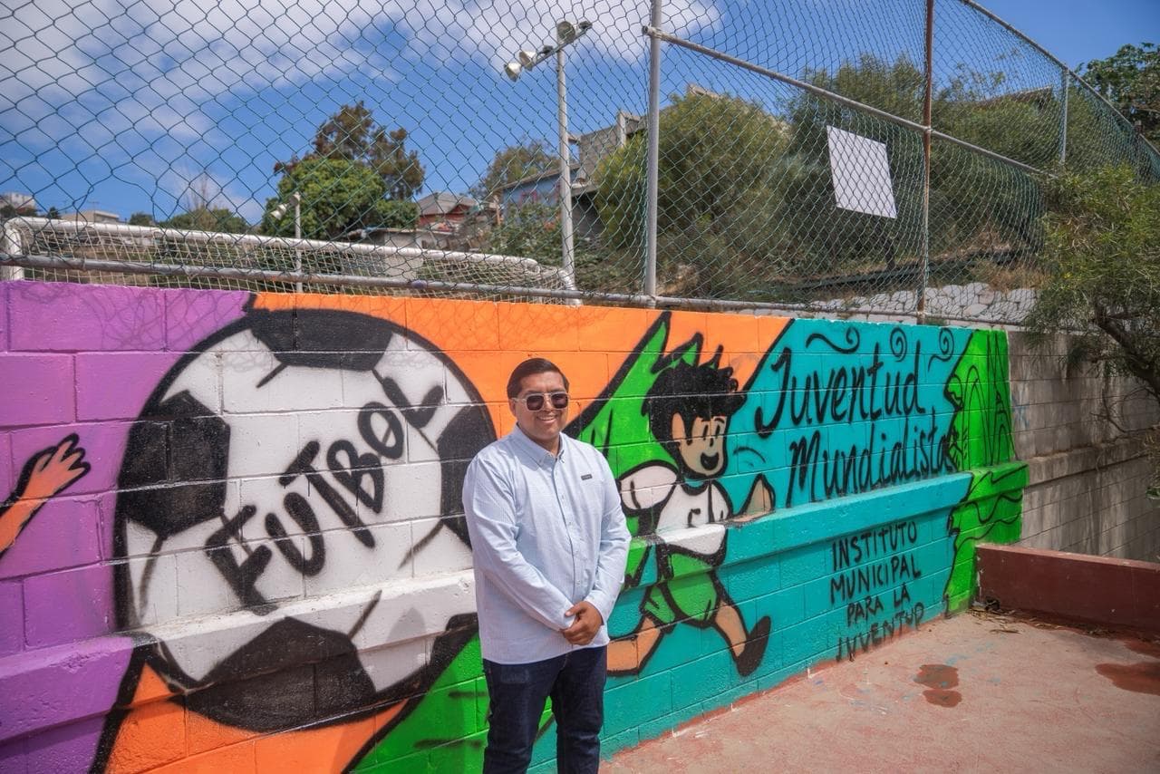 Mural colorido que promueve el fútbol y la juventud, con un joven posando frente a él en un espacio comunitario.