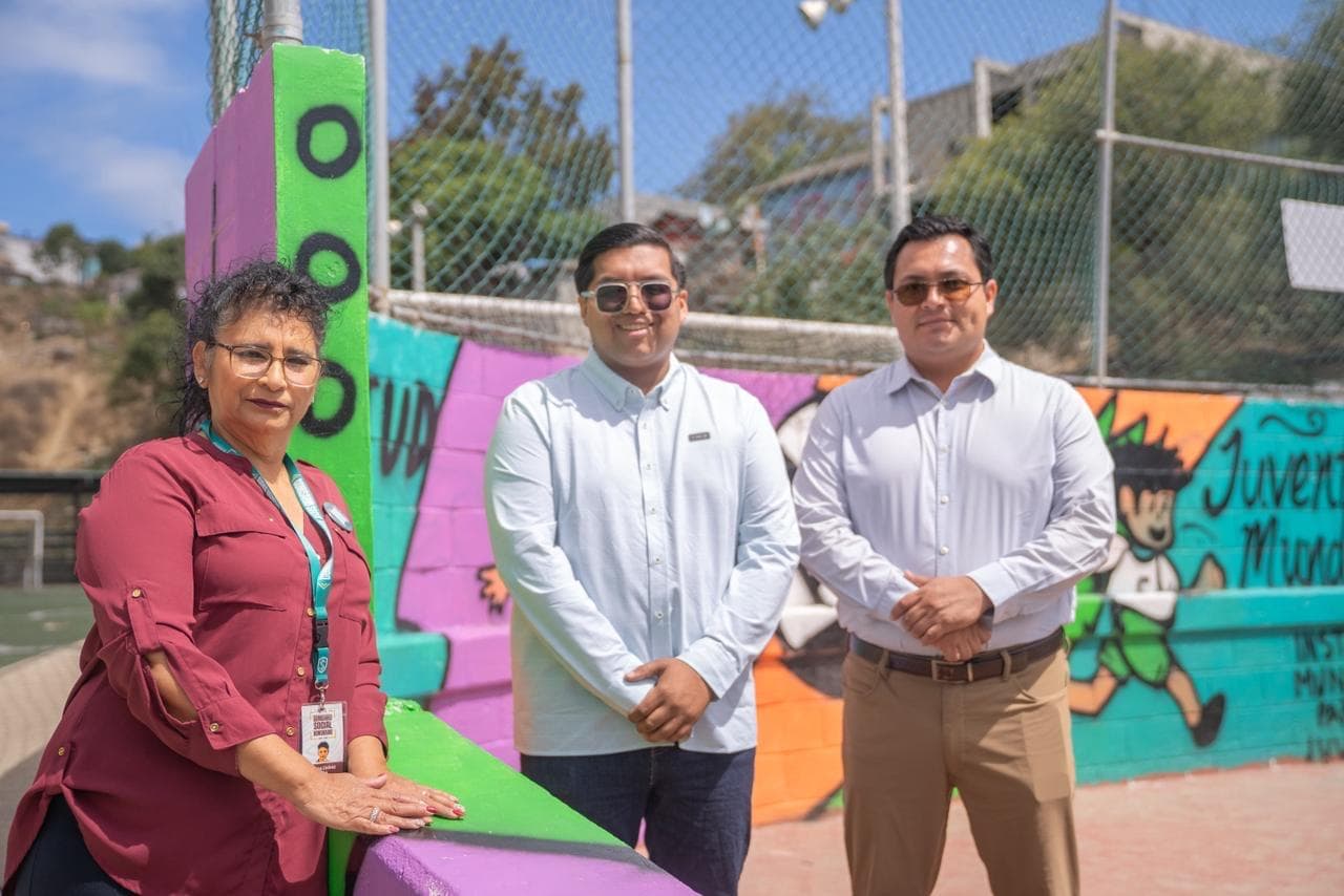 Tres personas posan frente a un mural colorido en una cancha deportiva, destacando la importancia del deporte juvenil en la comunidad.