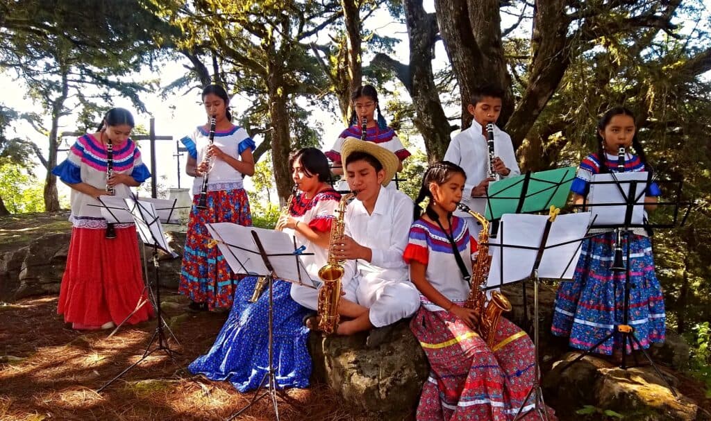 Niños y jóvenes tocando instrumentos musicales en un entorno natural, vestidos con trajes tradicionales, promoviendo la