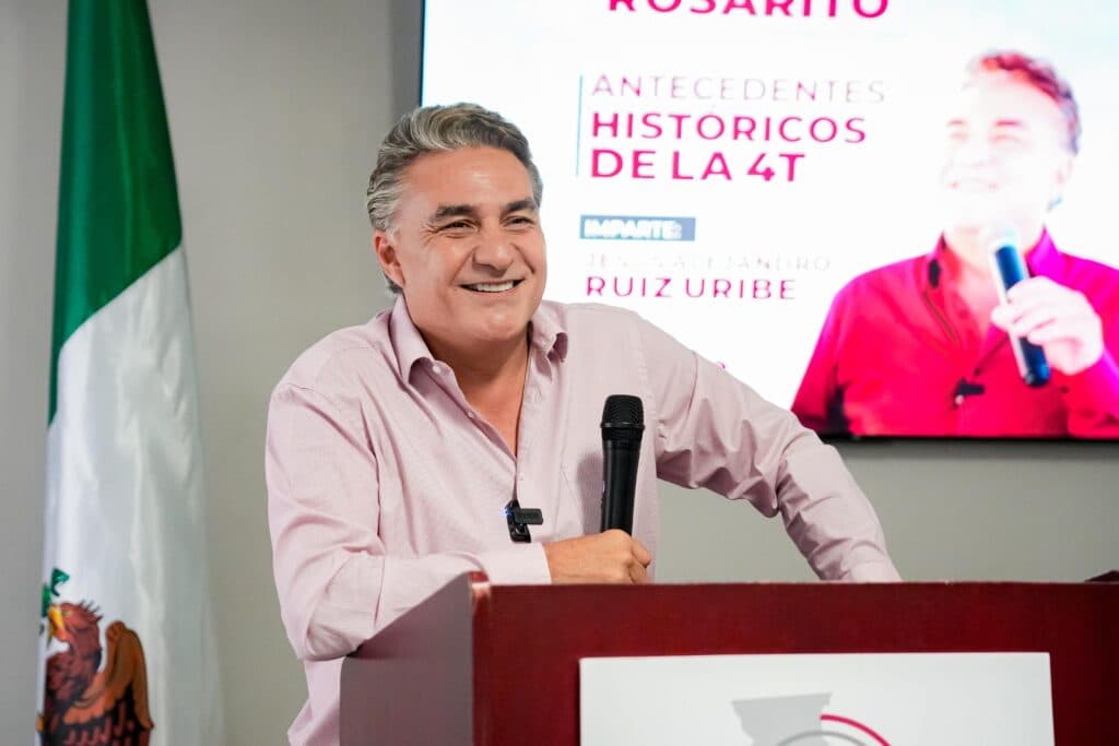 Jesús Ruiz Uribe sonríe mientras habla en un evento de MORENA en Casa Morena, Rosarito, con una bandera mexicana de fondo.