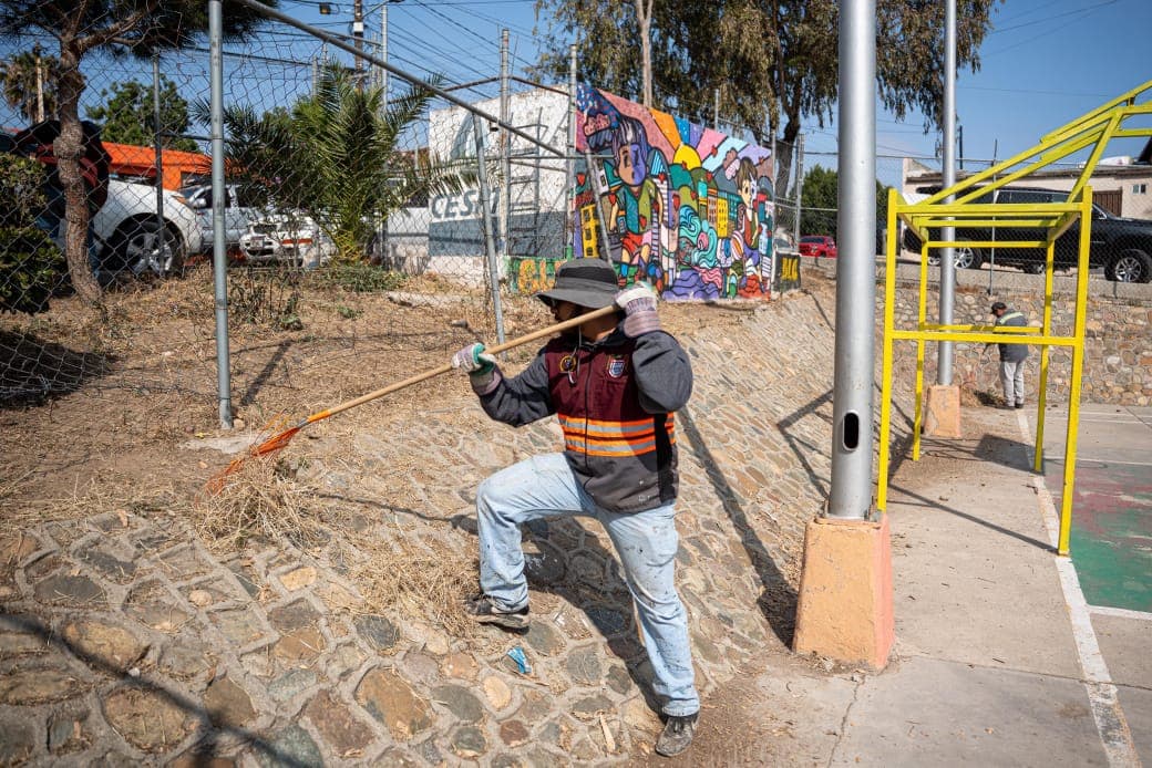 Trabajador de limpieza en un espacio público, utilizando una pala para retirar maleza junto a un mural colorido.