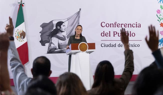 Claudia Sheinbaum Pardo durante la conferencia matutina en Palacio Nacional, con asistentes levantando la mano en señal de participación.
