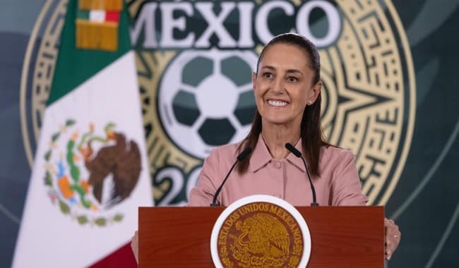 Claudia Sheinbaum sonríe mientras entrega boletos del Mundial 2026 en conferencia, con el logo del evento y la bandera de México de fondo.