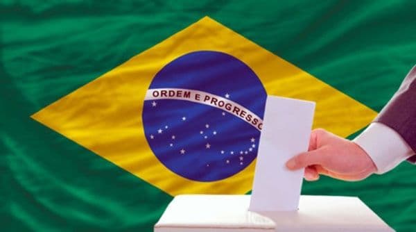Una mano depositando un voto en una urna, con la bandera de Brasil de fondo, simbolizando las elecciones presidenciales del país.