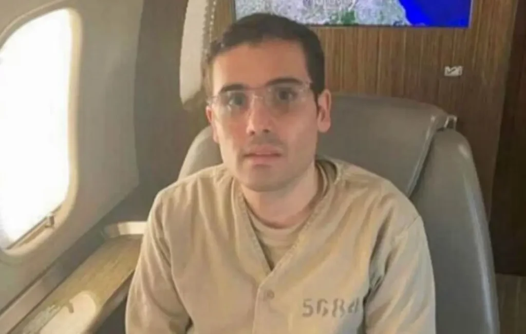 Ovidio Guzmán López, hijo de Joaquín "El Chapo", sentado en un avión, vestido con uniforme carcelario, en el contexto de su audiencia en EE. UU.